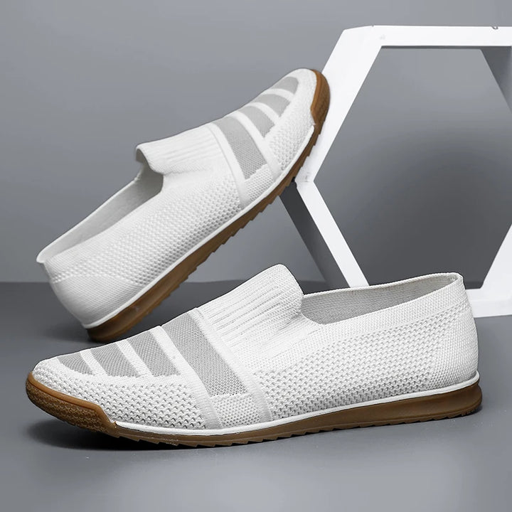 A pair of white slip-on men's casual shoes with textured knit material and brown rubber soles, offering a breathable loafers design, displayed against a gray background with a geometric white decor piece.