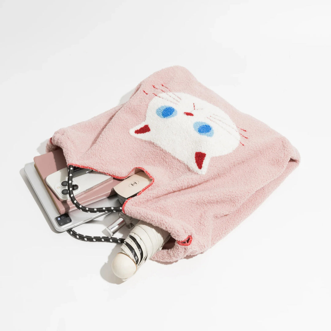A pink, fuzzy pouch crafted from lamb like fabric with a white cat face design is partially open, revealing a phone, a notebook, a pen, an umbrella, and a charging cable spilling out onto a white surface.