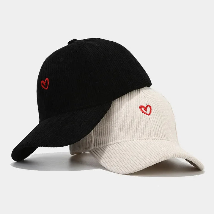 Two corduroy hats, one black and one off-white, each feature a small red heart embroidery on the front and are stacked atop each other against a plain white background.