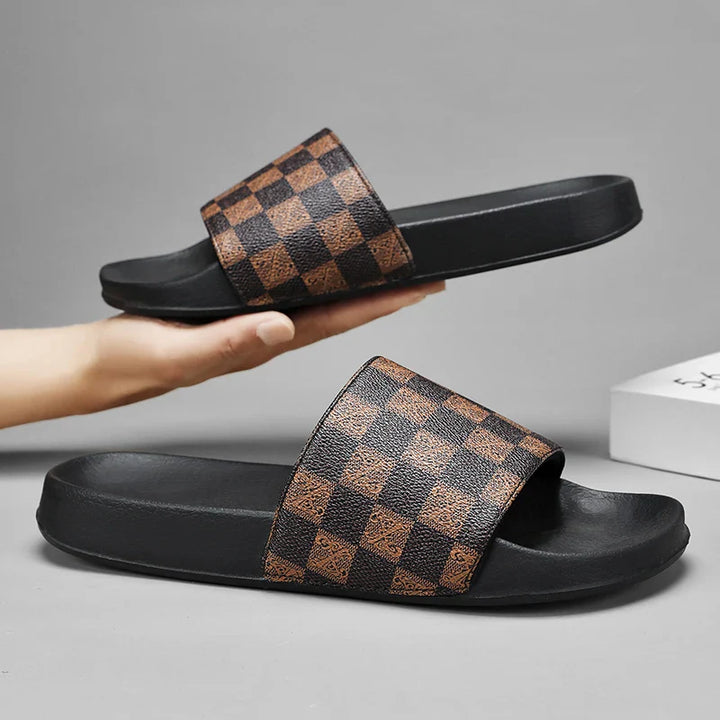 A pair of black checkered sandals with wide brown and black straps. One leather slip-on rests on a flat surface, while the other is held above it by a hand against a neutral gray background.
