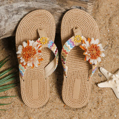 A pair of woven beige flip-flops with colorful zigzag-patterned straps and large orange and white fabric flowers, featuring an anti-slip sole—an ideal women's fashion accessory, placed on sandy ground next to a starfish and driftwood.