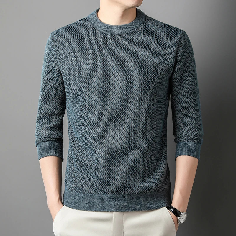 A person wears a relaxed fit sweater in textured teal with long sleeves and light-colored pants, standing against a gray background with hands in pockets. Their face is partially out of frame.