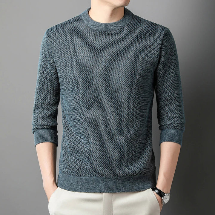 A person wears a relaxed fit sweater in textured teal with long sleeves and light-colored pants, standing against a gray background with hands in pockets. Their face is partially out of frame.