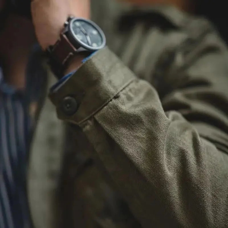 Close-up of a person wearing an olive green vintage military jacket with a buttoned cuff and a brown leather wristwatch. The person’s arm is raised against a dark, blurred background.
