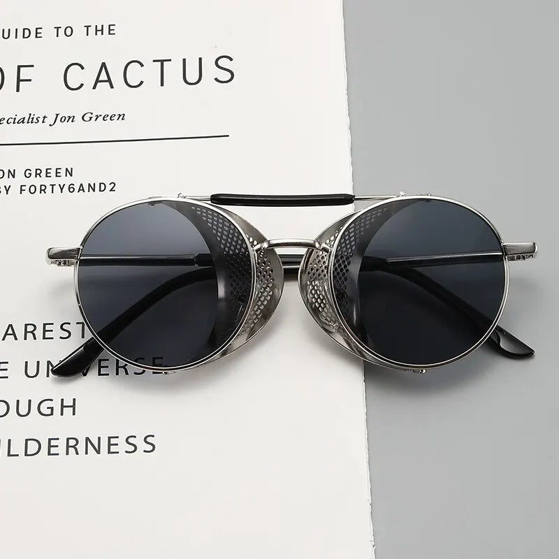 Round, silver-framed Steampunk sunglasses with dark lenses and perforated side shields rest on an open book or magazine with black text on crisp white pages.
