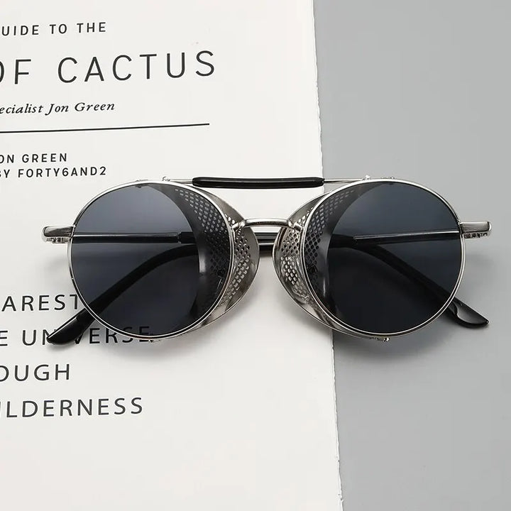 Round, silver-framed Steampunk sunglasses with dark lenses and perforated side shields rest on an open book or magazine with black text on crisp white pages.