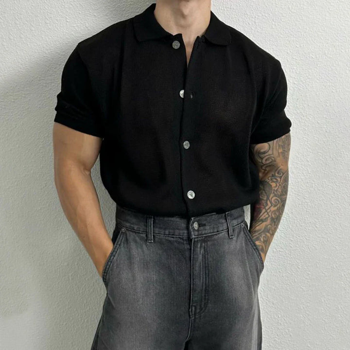 A person with a muscular build stands against a light-colored wall, wearing a black short-sleeve, button-up shirt and high-waisted, faded gray jeans. One arm features a tattoo sleeve, and both hands are in their pockets.