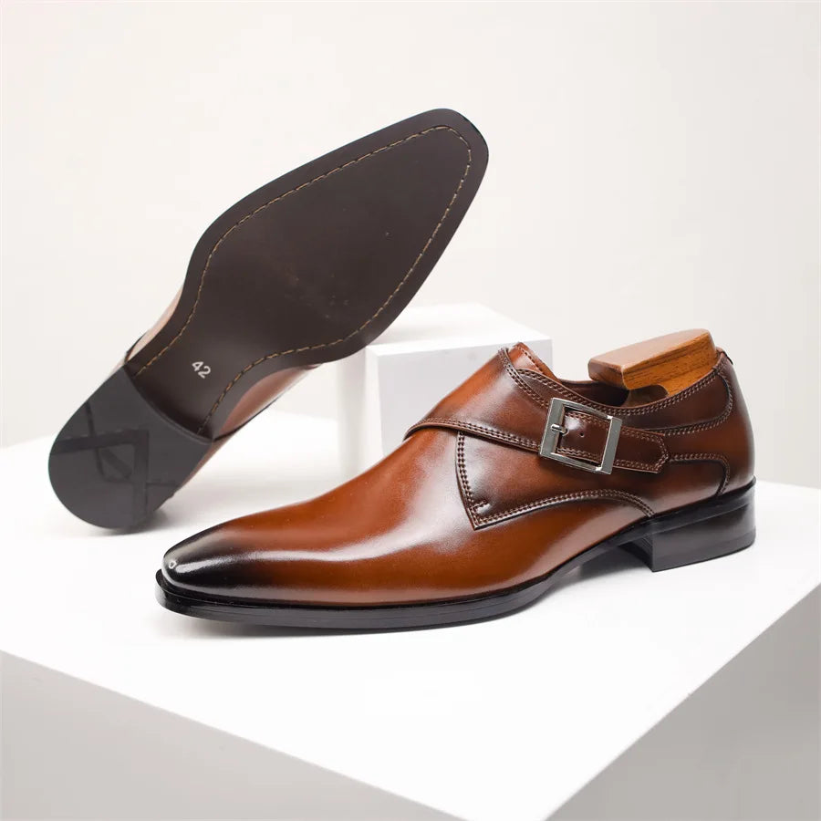 A pair of polished brown Italian leather monk strap shoes with a single side buckle and low heel, one shoe upright and the other showing the smooth black sole, displayed on white stands.