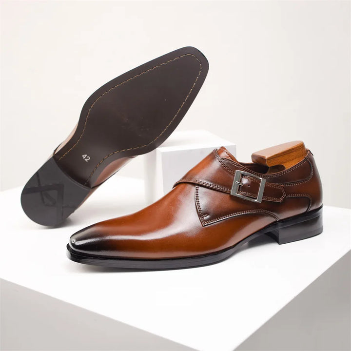 A pair of polished brown Italian leather monk strap shoes with a single side buckle and low heel, one shoe upright and the other showing the smooth black sole, displayed on white stands.