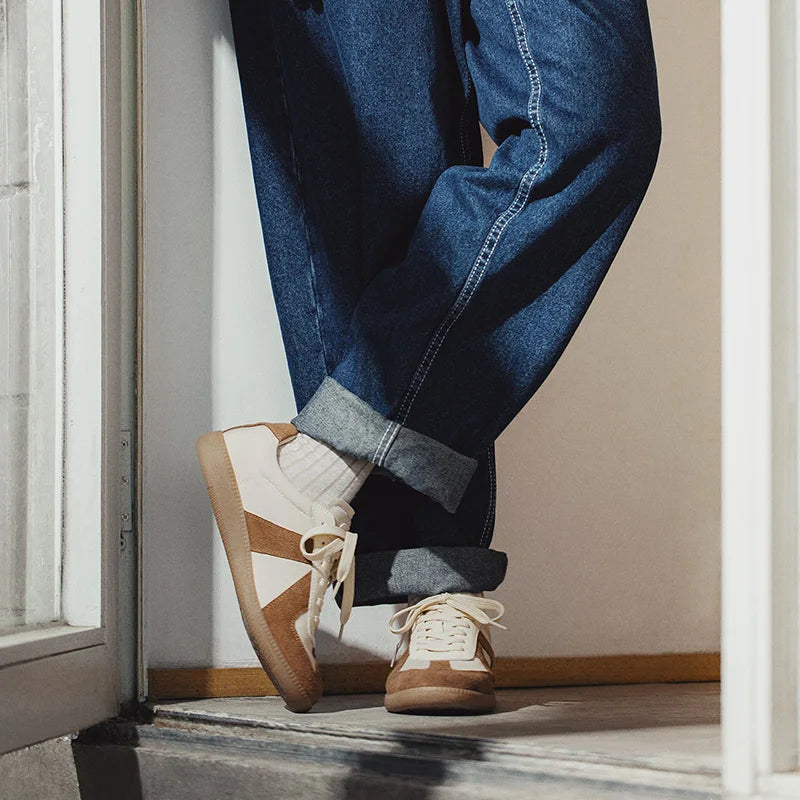 A person wearing rolled-up blue jeans, white socks, and lightweight breathable sneakers with premium cow suede and EVA midsole cushioning stands with legs crossed, leaning against a doorframe.