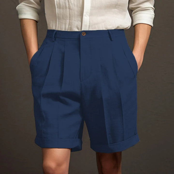 A person wearing a white button-up shirt with sleeves rolled up and high-waisted, breathable linen shorts stands with hands in pockets against a plain dark background—perfect for a relaxed summer look.
