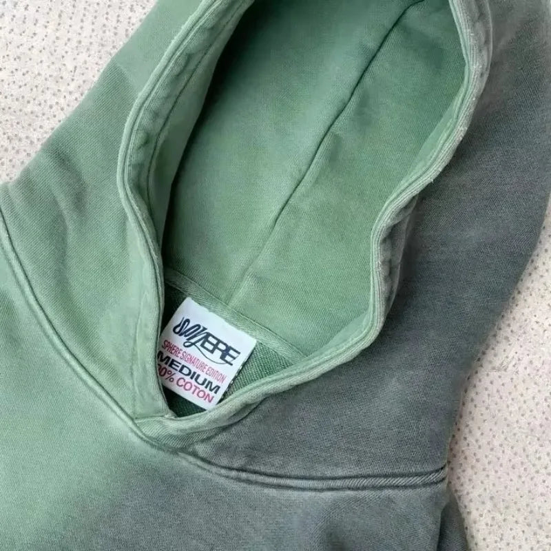 A green hooded sweatshirt made from breathable fabric with a visible SEE SEE MEDIUM 100% COTTON tag, lying on a textured light-colored surface.