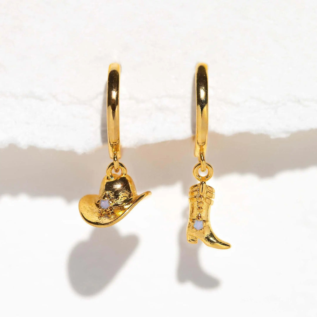 Two gold statement earrings, one with a dangling cowboy hat charm and the other with a cowboy boot charm, each accented with a small stone, set against a white background for an urban chic look.