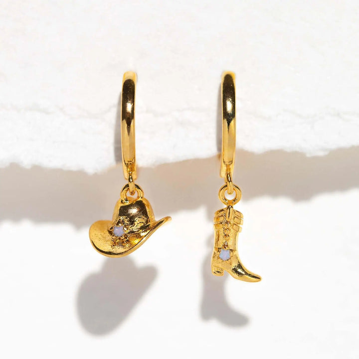 Two gold statement earrings, one with a dangling cowboy hat charm and the other with a cowboy boot charm, each accented with a small stone, set against a white background for an urban chic look.