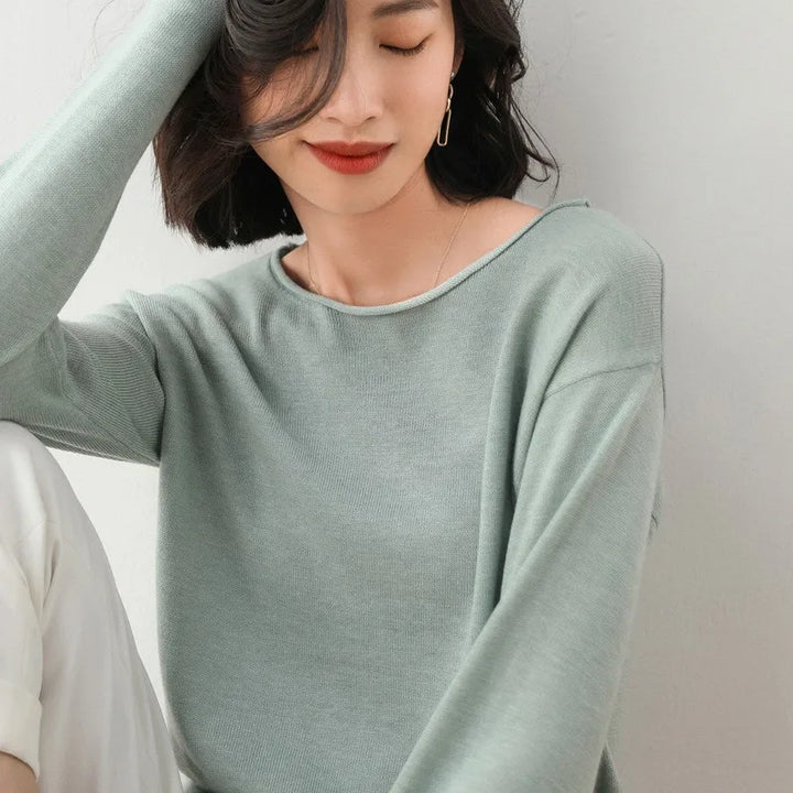 A woman with wavy dark hair and red lipstick wears a Japanese style light sage green relaxed fit cashmere blend long-sleeve top and white pants, sitting with one arm resting on her raised knee, smiling softly with her eyes partly closed.