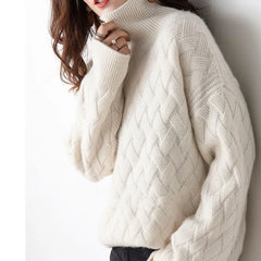 A person wearing a cozy, ivory-colored, ultra soft cable knit turtleneck sweater with long sleeves and textured patterns, paired with dark pants. The persons face is partially obscured by their hand.