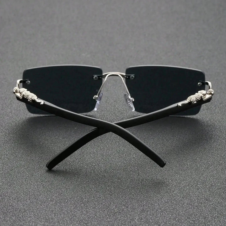Rimless black sunglasses with UV400 protection, dark lenses, and silver accents on the temples, displayed with the arms crossed on a textured dark surface.
