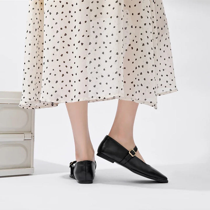 A person wearing a white, polka-dotted skirt and black Augusta Leather Ballet Flats with a buckle stands near a light-colored box, shown from the knees down against a white background.