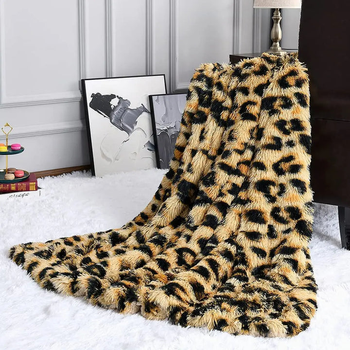 A plush throw in ultra-soft textile, boasting a chic leopard print blanket design, drapes over a dark chair on a white fluffy rug. Framed black and white artwork leans against the gray wall beside a lamp.