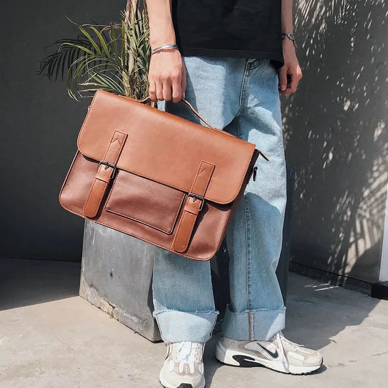 A person wearing light blue cuffed jeans, white sneakers, and a black shirt stands holding a brown premium leather briefcase with a spacious interior, with a potted plant in the background.