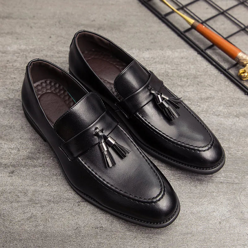 A pair of polished leather tassel loafers with decorative tassels on top, placed on a gray surface near a metal grid and a wooden-handled shoehorn.
