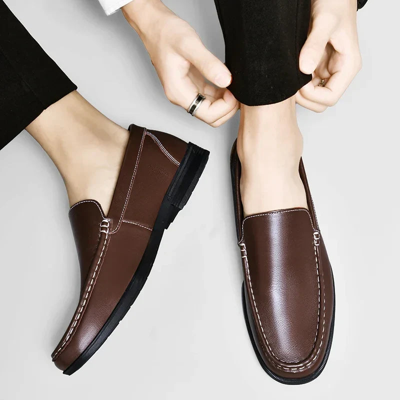 A person wearing black pants puts on a pair of classic slip-on loafers crafted from genuine leather, featuring white stitching and black soles.