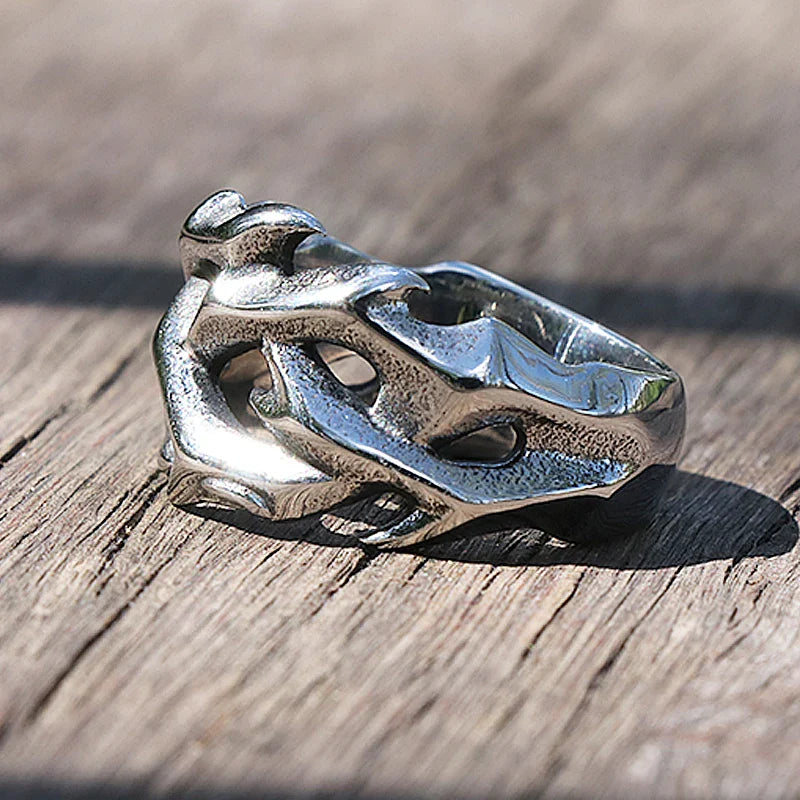 A silver streetwear ring with an abstract, sculpted design resembling intertwined shapes is placed on a wooden surface in natural light.