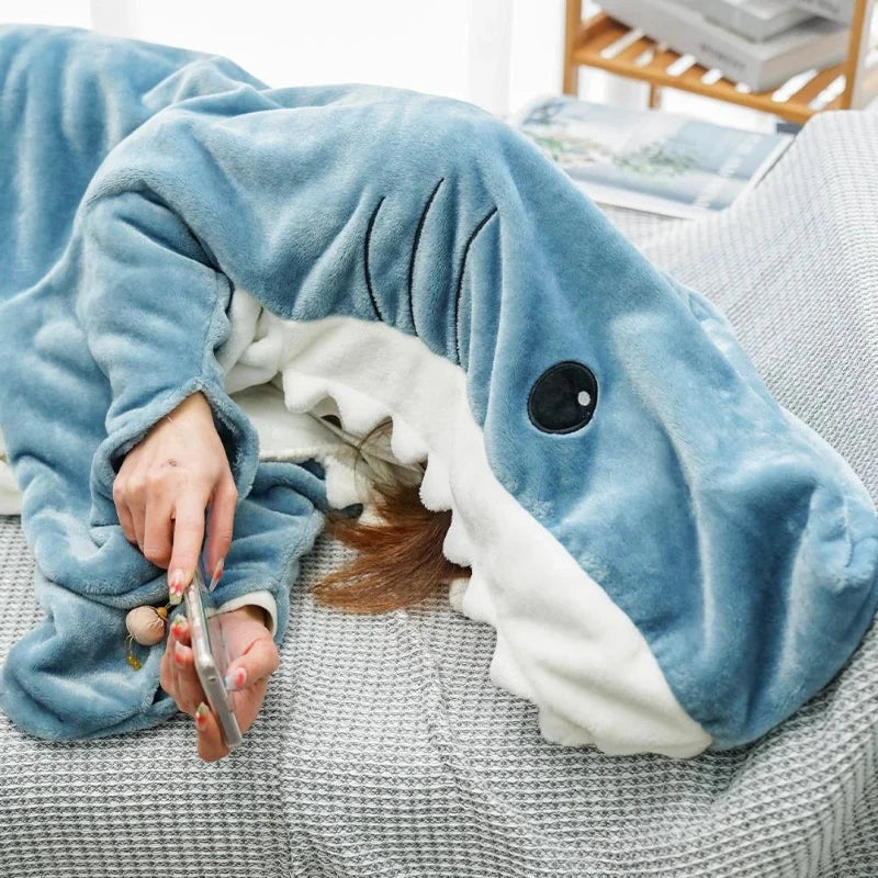 A person is lying on a couch covered by a plush blue and white shark sleeping bag, holding a smartphone in one hand. The sleeping bag covers most of their body and head, with only their hand and part of their face visible.