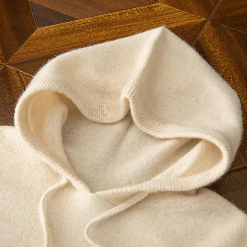 A close-up of a soft, cream-colored cashmere blend hooded sweater with drawstrings, lying flat on a wooden, geometric-patterned surface.