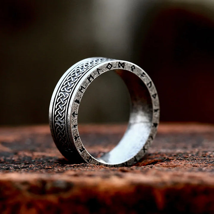 A durable metal Celtic knot ring with intricate patterns and ancient runic inscriptions stands upright on a rustic, textured surface against a blurred brown background.