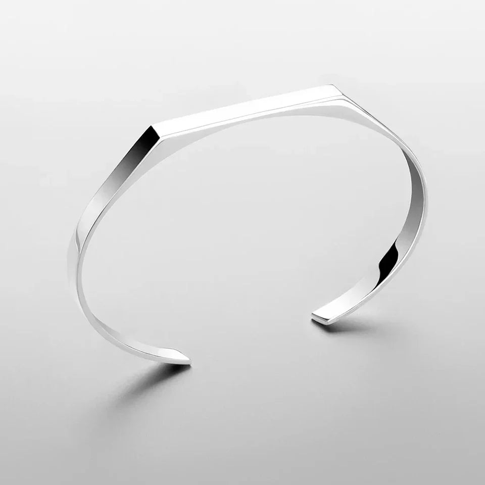 A minimalist cuff bracelet with a geometric, angular design and crafted from stainless steel is displayed on a light gray background, casting a subtle shadow.