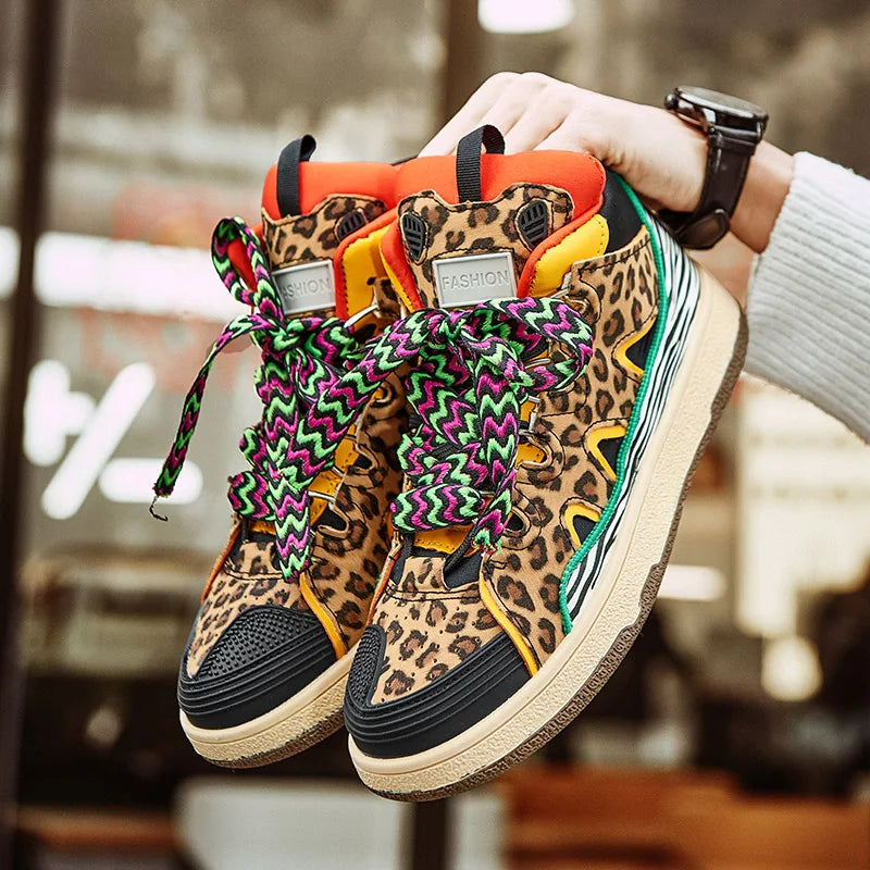 A person holds up a pair of lightweight, animal print shoes—colorful high-top sneakers featuring leopard print, bright orange and green accents, durable rubber soles, and patterned multicolored laces. The background is blurred.