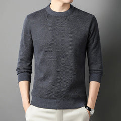 A person wearing a high-quality cotton sweater in dark gray with long sleeves and light-colored pants stands against a plain gray background, hands in pockets. The persons head is not fully visible.
