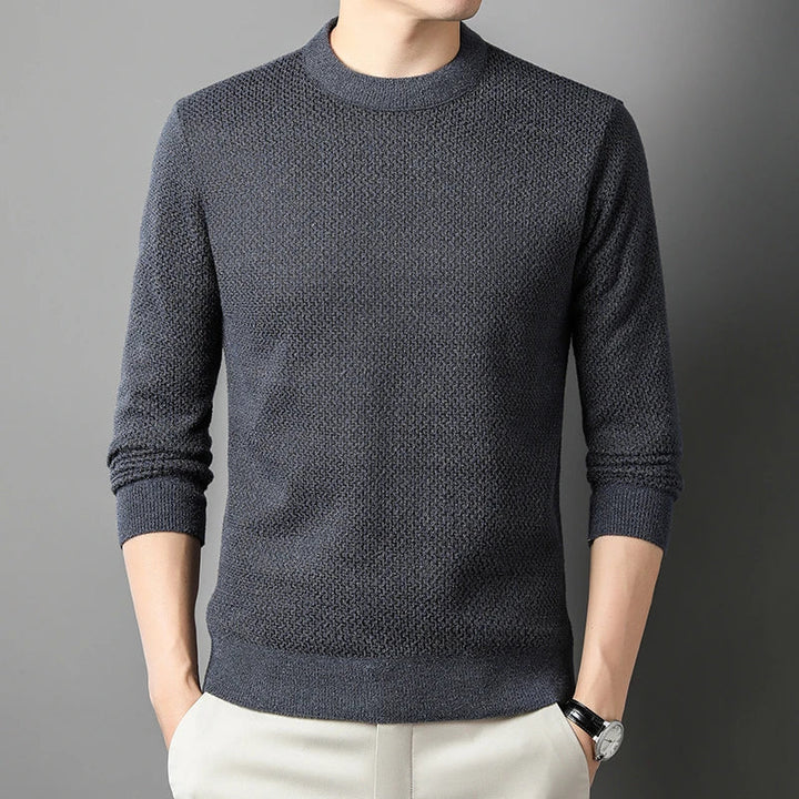 A person wearing a high-quality cotton sweater in dark gray with long sleeves and light-colored pants stands against a plain gray background, hands in pockets. The persons head is not fully visible.