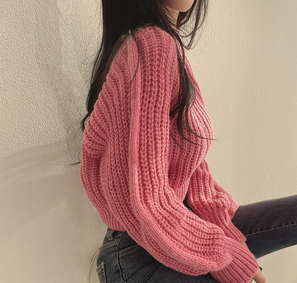 A woman with long dark hair sits sideways against a light-colored wall, wearing a chunky pink sweater made of soft knit material and dark jeans. The sunlight casts soft shadows on the wall.