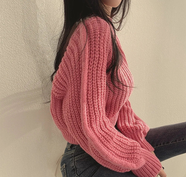 A woman with long dark hair sits sideways against a light-colored wall, wearing a chunky pink sweater made of soft knit material and dark jeans. The sunlight casts soft shadows on the wall.