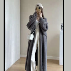 A person stands indoors taking a mirror selfie, wearing a long gray coat with a relaxed drape fit, white pants, a beige knit beanie, and a small white crossbody bag. Their face is partially covered by their phone.