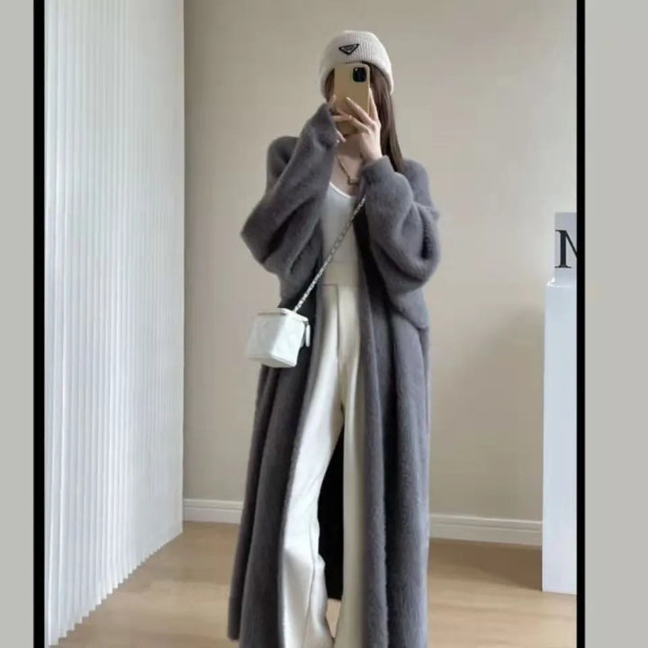 A person stands indoors taking a mirror selfie, wearing a long gray coat with a relaxed drape fit, white pants, a beige knit beanie, and a small white crossbody bag. Their face is partially covered by their phone.