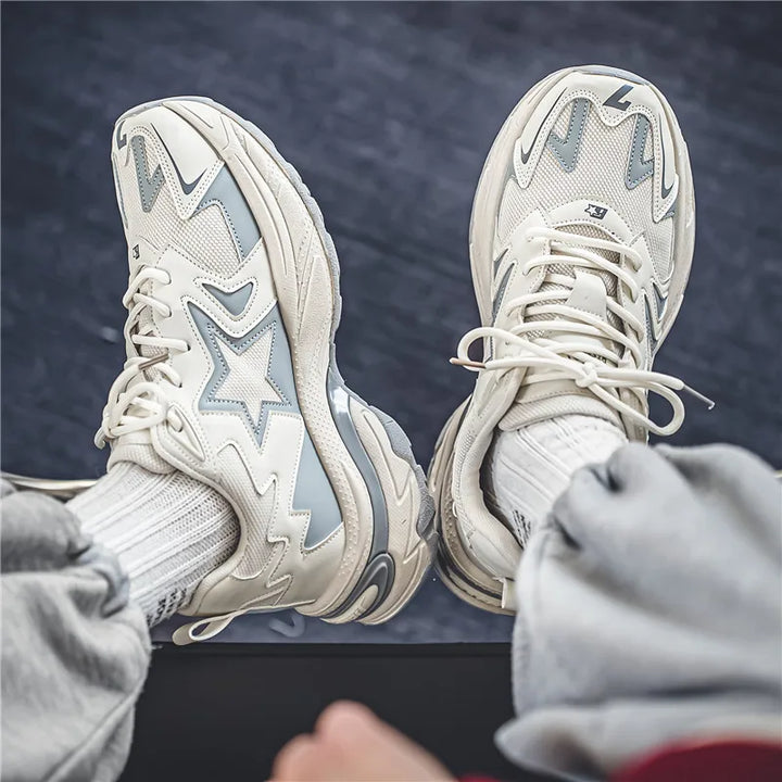 A person wearing light gray and white sneakers with star and wave designs, featuring a breathable mesh upper, white socks, and gray sweatpants sits with legs crossed on a dark blue carpeted floor.