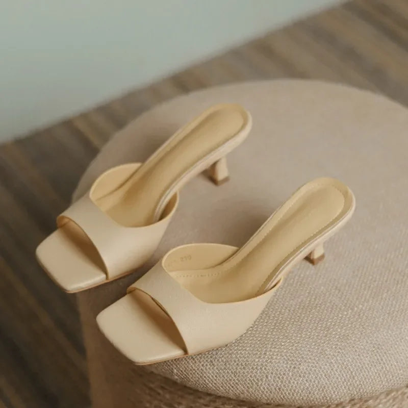 A pair of beige, open-toe, slip-on heeled sandals with genuine leather uppers and square toes rest on a round, textured ottoman.
