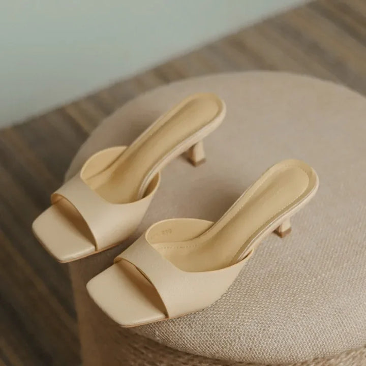 A pair of beige, open-toe, slip-on heeled sandals with genuine leather uppers and square toes rest on a round, textured ottoman.