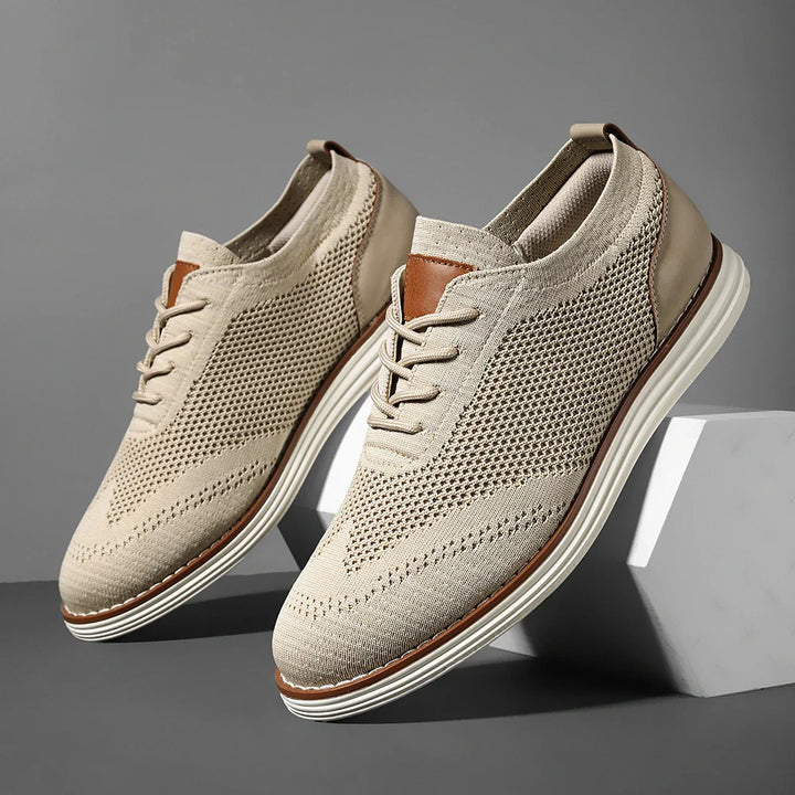 A pair of beige breathable knit sneakers with white soles and brown leather accents, displayed against a gray background with one shoe elevated on a white block for an effortlessly casual style.