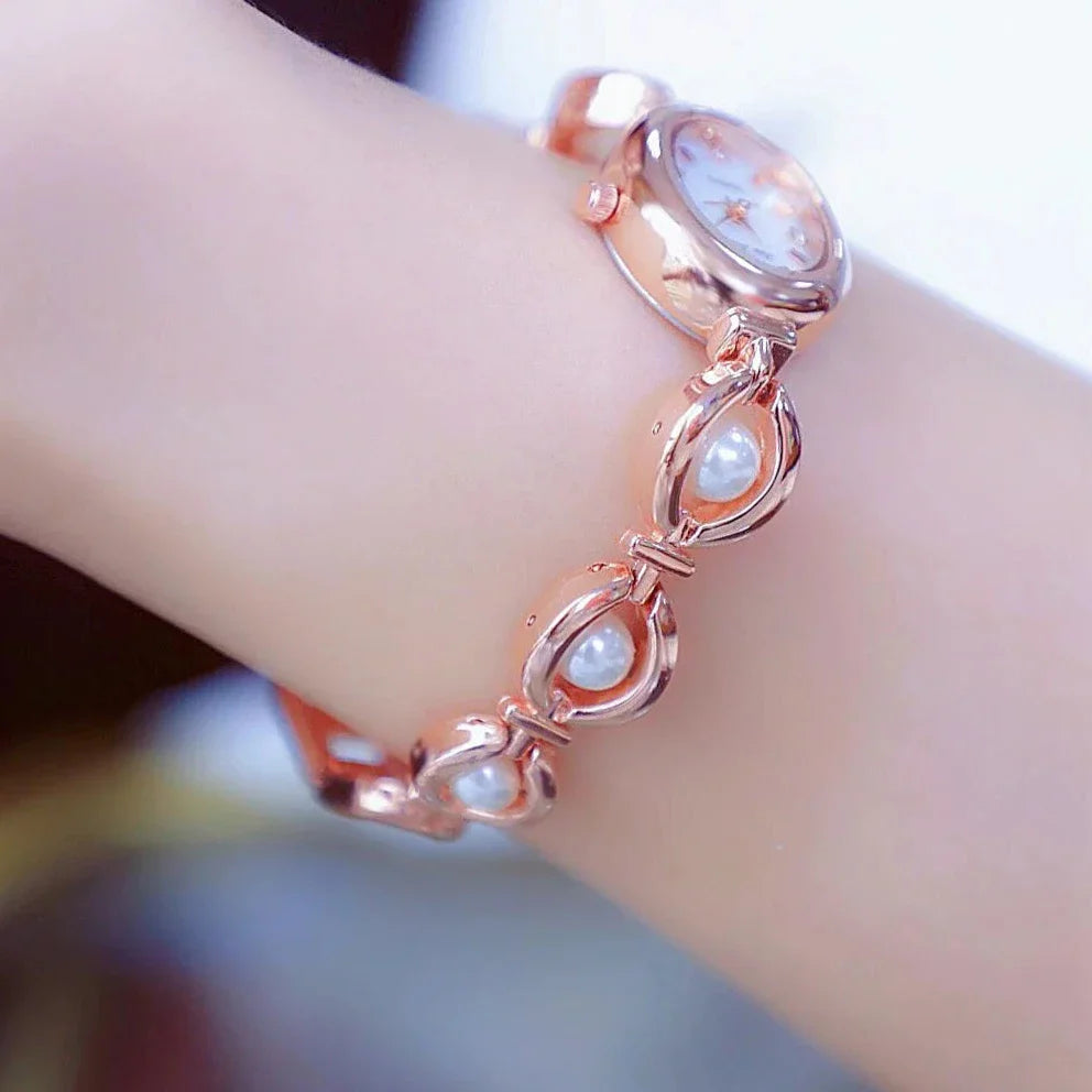 A rose gold wristwatch with a round face and a decorative band featuring leaf-shaped links and pearl accents, worn on a persons wrist.