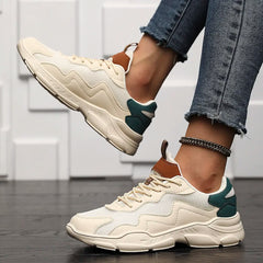 A person wearing cream-colored slip-on street shoes with green and brown accents, paired with frayed-hem skinny jeans and a silver anklet, stands on a dark wooden floor with a white textured wall in the background.