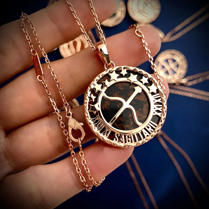 A hand holds a gold zodiac necklace featuring the Sagittarius symbol, surrounded by stars and the word SAGITTARIUS at the bottom. Several astrology jewelry pieces with a copper finish are blurred in the background.
