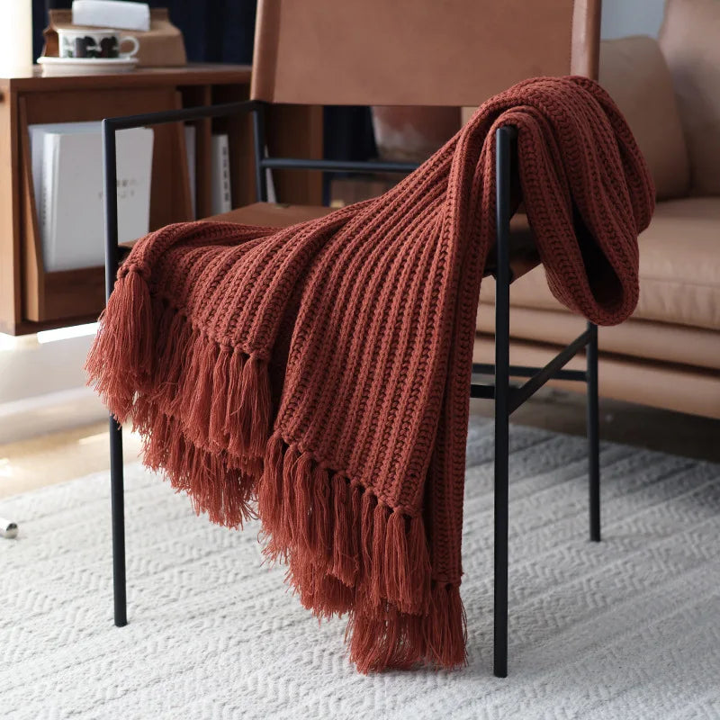 A rust-colored chunky weave throw with thick tassels drapes over a modern black metal chair, placed on a light textured rug near a tan leather sofa in a cozy living room setting.