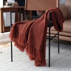 A rust-colored chunky weave throw with thick tassels drapes over a modern black metal chair, placed on a light textured rug near a tan leather sofa in a cozy living room setting.