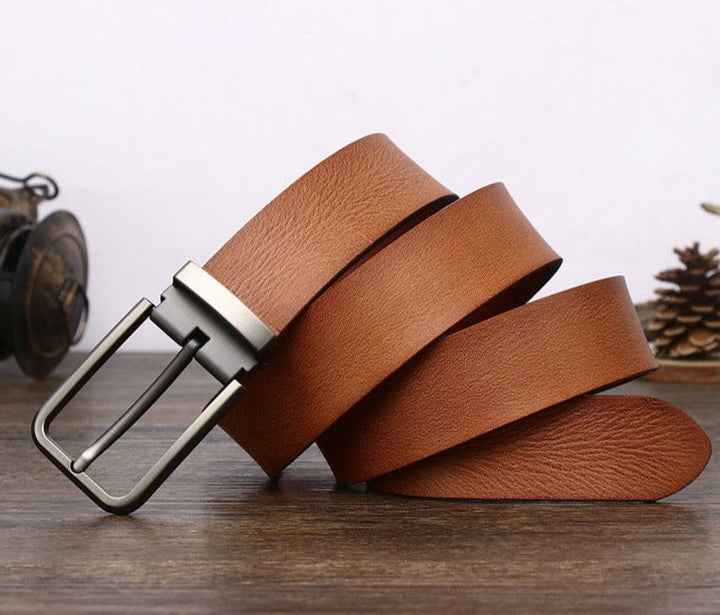 A brown cowhide belt with a silver buckle is coiled on a wooden surface. A pine cone and a vintage camera are blurred in the background.