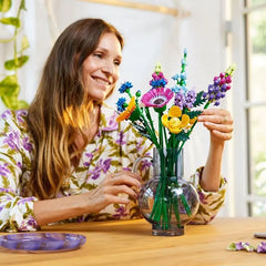 A smiling woman arranges a Wildflower Bouquet of colorful LEGO flowers in a clear glass vase while sitting at a wooden table, wearing a floral shirt, with bright natural light coming from a window behind her.