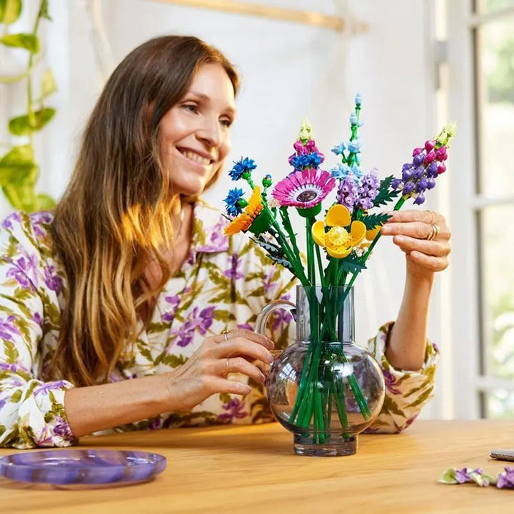 A smiling woman arranges a Wildflower Bouquet of colorful LEGO flowers in a clear glass vase while sitting at a wooden table, wearing a floral shirt, with bright natural light coming from a window behind her.